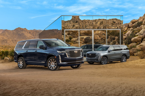 Two luxury SUVs parked on a desert landscape, with a modern glass building reflecting rocky hills. Clear blue sky above, creating a sleek, adventurous tone.